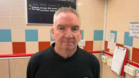 Stuart Devine es un hombre mayor con cabello gris parado en una tienda de chips de Ashvale y con una camiseta de Ashvale dibujada a lápiz a la espalda. En la pared detrás de él hay azulejos rojos, crema y azules estilo madera contrachapada, ranuras y hojas informativas colgadas en otras paredes visibles.